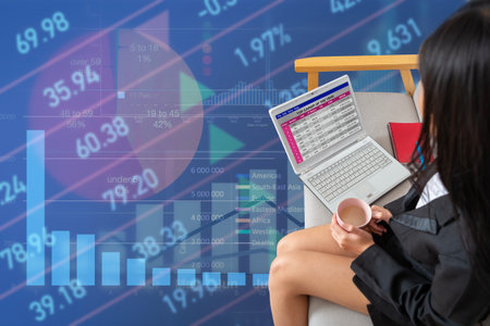 Businesswoman drinks a cup of coffee and works relaxed on laptop with stock data and financial...