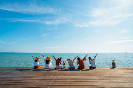 Photos from the back of Asian women lined up on a terrace by the ocean in the morning. They raise one arm oblique, both sides of the left and right. Feelings of fun and happiness. Copy space on top.の写真素材