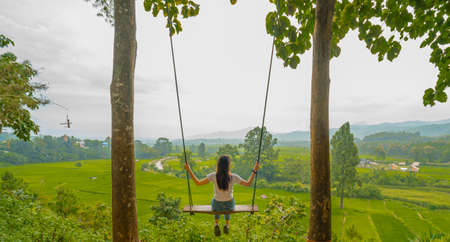 Rear shot of an Asian woman sitting on a swing on a high cliff. There are views of the vast rice fields, greenery, and houses in the countryside. feeling relax and fun. There is a copy space on top.の写真素材