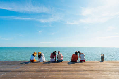 Photos from the back of Asian women wearing colorful colored clothes sitting on the balcony by the sea. They tilted their heads together in pairs. Feelings of love, friendship, and happiness.の写真素材