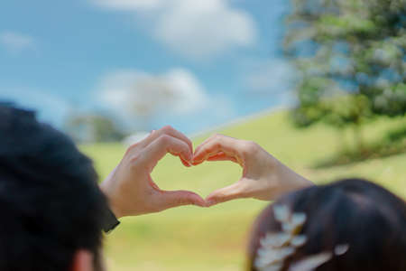 Photos from the back of a man and a woman raise a hand above the head and pose in heart shape. There is a blurred backdrop of field and blue sky. Feelings of love and happiness. Copy space on top.の写真素材