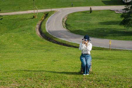 A woman with a white long sleeve shirt sits on a stump in a lush grassy hill. She was holding a camera taking pictures in front of her. The backdrop is curved concrete roads and intersections.の写真素材
