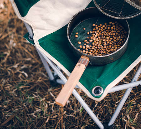 Close-up photo of a dark brown color of coffee bean sieve with the wooden handle placed on a foldable green chair. The floor is dry grass. Feeling relax and fresh. There is a copy space on left.の写真素材