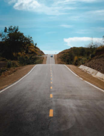 Blur background of gray asphalt road in a peaceful rural area on a sunny day and clear blue sky clouds. There are white border and yellow central line. The roadside is brown, grass, and green trees.の写真素材