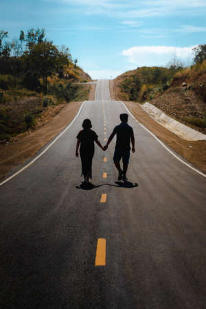 Men and women walk hand in hand on asphalt road in a rural area on a sunny day and clear blue sky clouds. There are a white border and a yellow central line. The roadside is brown and green trees.の写真素材