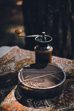 Close-up photo of a coffee bean sieve and a wooden coffee grinder placed on a bohemian brown table cover. There is sunlight shining on the object. Feeling relax and fresh.の写真素材