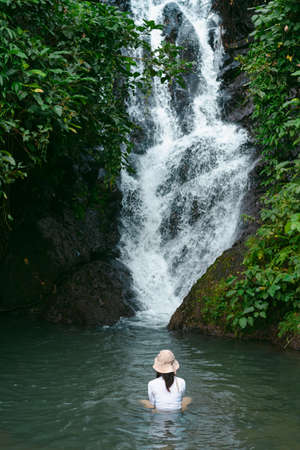 A young woman wearing a white t-shirt and hat, sitting with her back turned on the rock at waterfall in rainy season, Thailand. Feeling fresh, relaxed, and freedom. Idea for adventure nature tourism.の写真素材