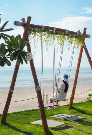 Picture from the back of a woman sitting on a swing that is tied to the bar in the morning. The top has ivy and the lower has green grass. Blurred backdrop is a blue ocean with glitter and clear sky.の写真素材