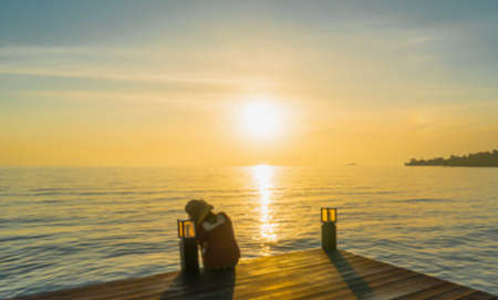 A woman sat back on balcony looking at the rising sun over the edge of the water early in morning. The orange and blue light of sky blended perfectly. There was a glittering reflection on a calm sea.の写真素材