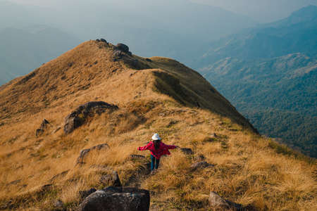 A woman with a red long-sleeved shirt and white hat walk and open arms on golden dry grasslands at the ridge of Mulayit Taung, Myanmar. There are overlapping hills and many rocks. There is copy space.の写真素材