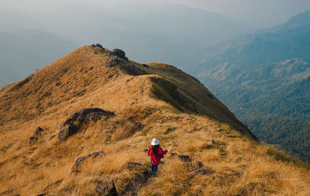 A woman with a red long-sleeved shirt and a white hat walks on golden dry grasslands at the ridge of Mulayit Taung, Myanmar. There are overlapping hills and many rocks. There is copy space on top.の写真素材