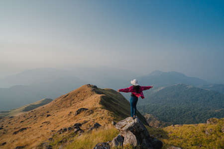 A woman with a red long-sleeved shirt and white hat stand and open arms on the stone at the ridge of Mulayit Taung, Myanmar. There are overlapping hills and golden dry grasslands. There is copy space.の写真素材