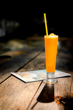 Isolate of a glass cold orange smoothie juice place on the wooden table with brown color. Menu blurred backdrop. Sunlight shining causing shadows. Feeling refreshing. Summer and vacation concept.の写真素材