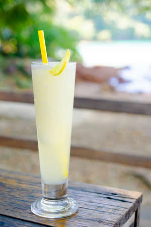 Isolate of a glass cold lemon smoothie juice place on the wooden table with brown color. Blur backdrop is sea, sky, beach, and trees. Feeling refreshing and Sour taste. Summer and vacation concept.の写真素材