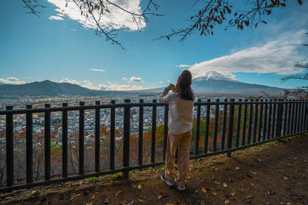Photos from the back of an Asian woman standing by the side of the Chureito Pagoda, Japan. There is Mount Fuji with snow at the top with views of the city near the volcano in the autumn.の写真素材