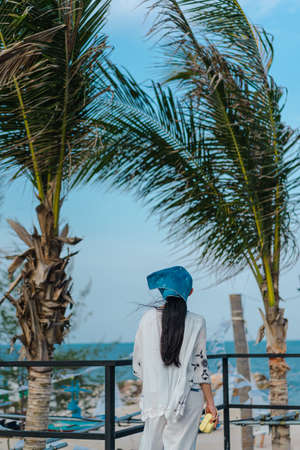 Photo from the back of woman stands on terrace with blurred background of coconut trees and blue ocean in evening. There is windy day with clear blue sky. Feeling fresh  and cool. There is copy space.の写真素材