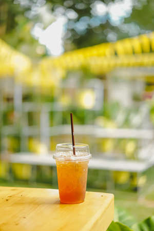 Close-up of cool peach tea in clear plastic glass on a wooden table on a sunny day. The blurred background of yellow, white, and green color with bokeh in a soft tone. There is a copy space on top.の写真素材