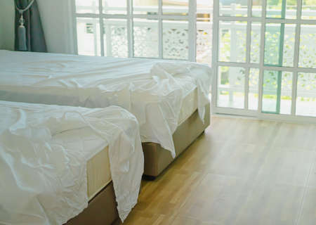 Double bed with wrinkled white sheets in the hotel. There are clear glass and straight edges in the morning. The floor is a simple light brown wood grain. Idea for unclean with copy space.の写真素材