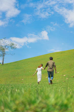 A photo from the back of a man and woman holding hands on a green grassy slope in soft tone. They are walking to the top of the hill on a clear blue sky with white clouds. There is copy space on top.の写真素材