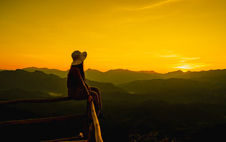 Picture from the back of a woman sitting on wooden porch extending into a high mountain cliff. The sun is setting on the mountain and there is a beautiful warm orange light. The traveling background.の写真素材
