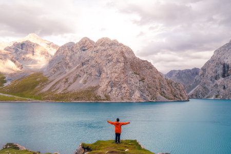 A woman stands on a rock with lush green grass by the Tientang Hu lake, Xinjiang, China. There are many mountains behind. The soft cool light shines on the still and shimmering blue water surface.の写真素材