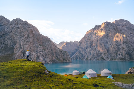 The front view of Tientang Hu lake, Xinjiang, China. There are many mountains behind. There are many tents on the green lawn by the water. There is a woman in a white jacket standing on a cliff.の写真素材