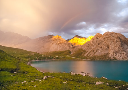 The front view of a lake. There are many mountains behind. The sun was setting with soft yellow light. There is a lush green lawn and a rainbow with mist on a cloudy day.の写真素材