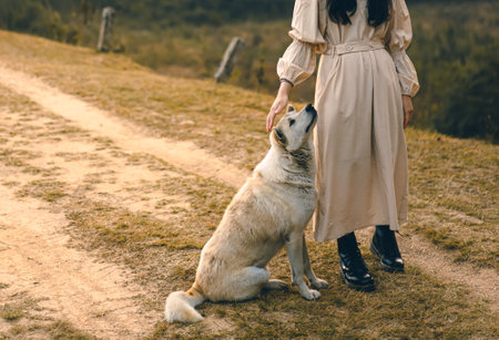 A white dog sits and is being petted by a woman next to him. A lady in a long skirt and black boots...