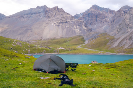 Front view of a grey tent with black camping chairs beside Tiantang Lake, Xinjiang, China. There are mountains in the background on a cloudy day. A dog is lying on its back on the green grass.の写真素材