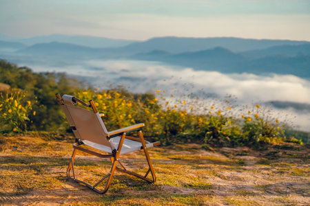A white camping chair on the ground with a misty mountain view on a cold day in the morning. There are yellow flower bushes on green grass in soft tone. Idia for outdoor background with copy space.の写真素材