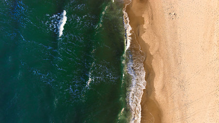 Aerial drone view of the calm Baltic Sea near Sarbinowo, Poland. Gentle waves wash over golden sand under clear spring sunlight, creating a peaceful coastal scene.の写真素材