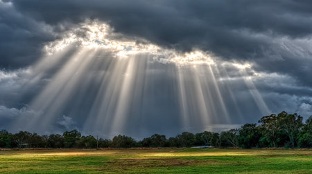 This stunning image captures the dramatic interplay of clouds and sunlight, with rays piercing through dark stormy skies over a lush green field.の素材