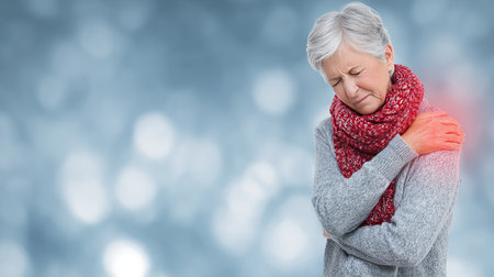 A senior woman with gray hair, wearing a cozy red scarf, expresses discomfort as she holds her shoulder. The blurred background creates a calm atmosphere, highlighting her health concerns.の素材