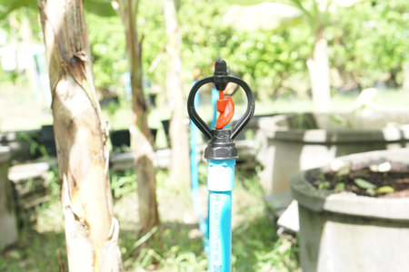 A close-up view of a colorful garden sprinkler connected to an irrigation system. Lush plants surround the area, showcasing effective watering technology for gardens.の素材