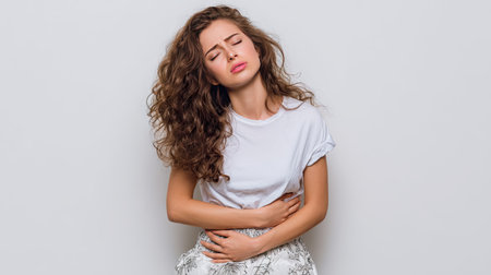 A young woman with curly hair shows discomfort while holding her stomach. Her expression reflects pain and concern, set against a plain background.の素材