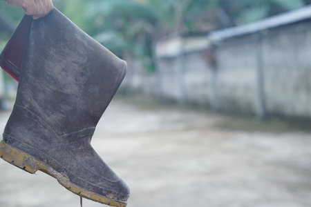 A close-up view of a muddy rubber boot held by a hand, set against a faded backdrop of a tranquil outdoor path, symbolizing farming life and hard work.の素材