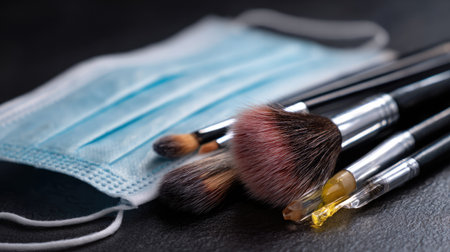 A collection of makeup brushes and a medical face mask arranged artistically on a dark surface, representing the intersection of beauty and hygiene.の素材