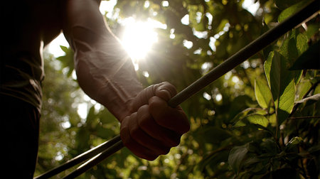 A close-up view of a strong hand gripping a metal rod, illuminated by sunlight filtering through vibrant green foliage, showcasing power and determination in nature.の素材