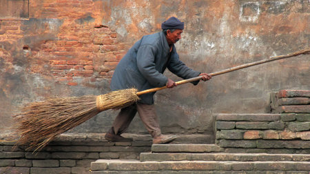 An elderly man wearing a coat sweeps steps with a traditional broom, surrounded by rustic brick walls, capturing everyday life and cultural heritage.の素材