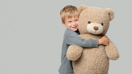 A joyful young boy embraces a large teddy bear, radiating warmth and innocence against a neutral backdrop. Perfect representation of childhood joy and affection.の素材