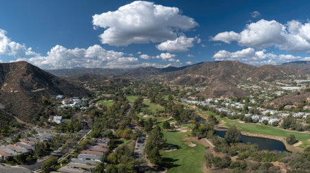 Experience the breathtaking aerial view of a tranquil golf course nestled in the foothills, showcasing vibrant greenery and majestic mountains beneath stunning clouds.の素材