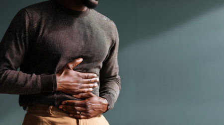 A man in a textured dark sweater gently holds his stomach as he stands against a softly lit background, reflecting a mood of introspection and health awareness.の素材
