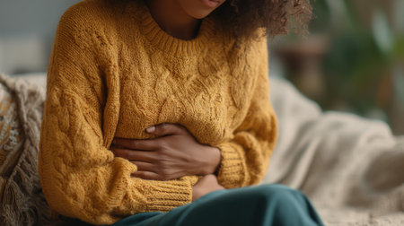 A young woman in a cozy yellow sweater sits on a couch at home, holding her stomach with a concerned expression, reflecting discomfort and unease.の素材