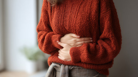 A woman in a cozy red sweater stands indoors, gently holding her stomach, conveying discomfort while surrounded by soft light and a calm atmosphere.の素材
