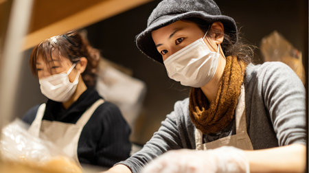 Two women engaged in food preparation in a cozy kitchen setting. Both are wearing masks and aprons to ensure hygiene and safety while working together.の素材