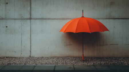 A vibrant orange umbrella stands alone against a smooth concrete wall, creating a striking visual contrast with the minimalist gravel surface below.の素材