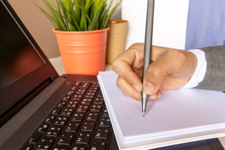 Human hand with the pen writing on the notebook that align over the laptop with the little tree and coffee cup and calendar.の写真素材