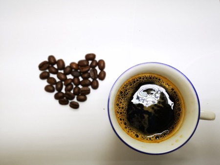 Top view of hot black coffee in white ceramic cup and coffee beans heart on white groundの写真素材