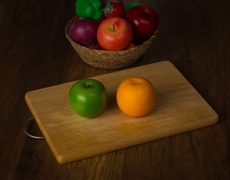 Green apple and orange on chopping block and fruits in a basket on desk backgroundの写真素材