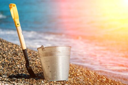shovel with a yellow plastic handle and a galvanized bucket on a pebble beach with the waves rolling in background sea with blue sky. Close up. Copy space. Travel, relax or loneliness concept.の写真素材